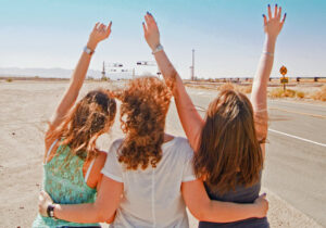 Drei Frauen mit erhobenen Armen von hinten auf einer Wüstenstraße, die mittlere mit lockigem rotem Haar