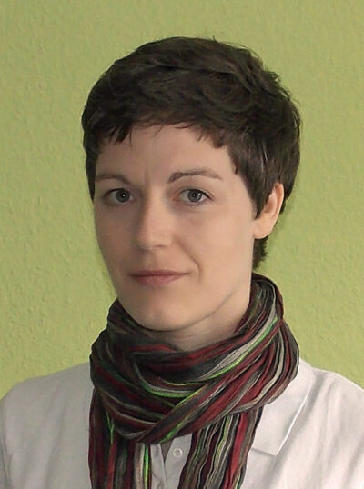 Marlen Weller-Menzel mit kurzem braunem Haar und buntem Schal, lächelt vor grüner Wand