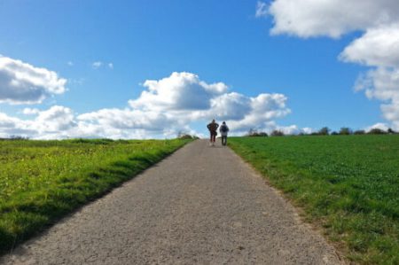 Zwei Menschen gehen gemeinsam auf einem geraden Weg durch grüne Felder unter blauem Himmel mit weißen Wolken