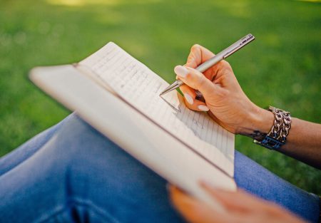 Hand mit Armband schreibt mit Stift in ein Notizbuch, Person sitzt im Freien auf grünem Rasen